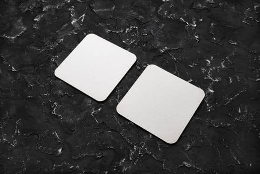 Two blank square beer coasters on black plaster background.