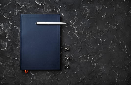 Blank blue notebook with pen on black plaster background. Copy space. Flat lay.