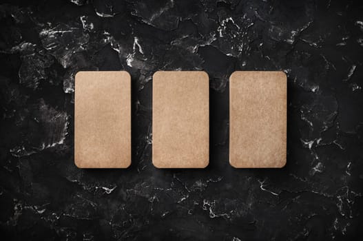 Three blank kraft business cards on black plaster background. Branding mockup. Top view with copy space. Flat lay.