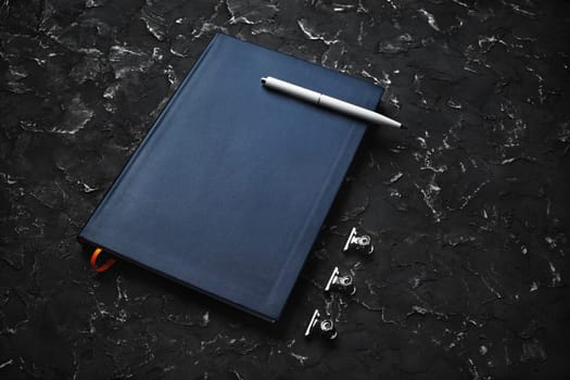 Blank blue notepad, pen and clips on black plaster background.