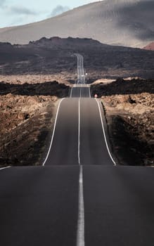 Endless road on a volcano in Timanfaya National Park in Lanzarote in the Canary Islands with a continuous line, black volcanic rocks on the side and volcanoes in mist in background