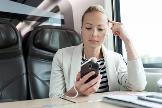 Caucasian businesswoman communicating on cellphone using headphone set while traveling by train in business class seat.