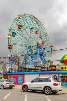 Wonder Wheel Ferris Wheel at Luna Park Amusement Park with stores and vending machines in front, Coney island, Brooklyn, New York City during winter day with cloudy sky, Cars parked next to the street in front, vertical