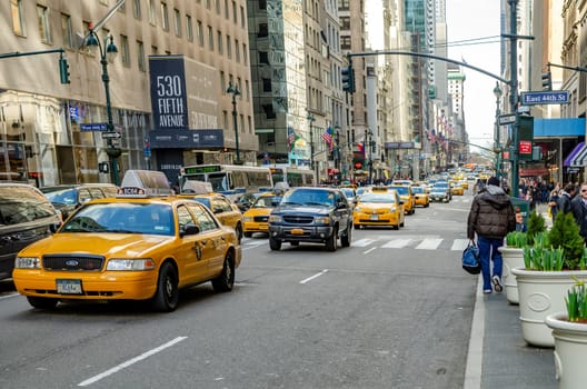 Fifth Avenue Street with lots of Yellow Taxi and cars passing by, lots of People walking on the sidewalk, Different Stores and office Buildings, American flags hanging down the Buildings, New York City during winter day with cloudy sky, horizontal