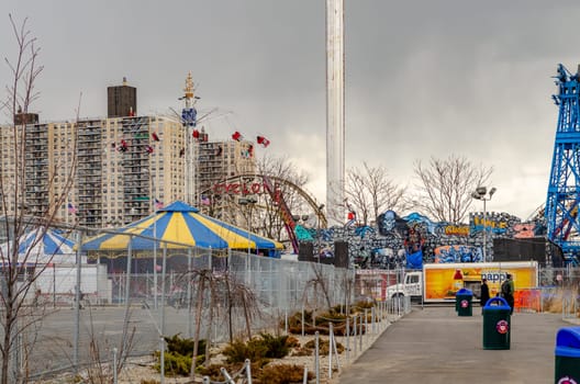 Rides at Luna Park Amusement Park, Coney island, Brooklyn with Fence, footpath and trash can in front, New York City during winter day with cloudy sky, Residential Building in the background, horizontal