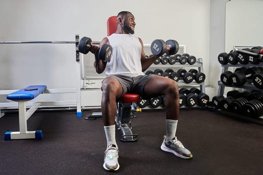 An African American man shakes his biceps while sitting on a bench in the gym. High quality photo