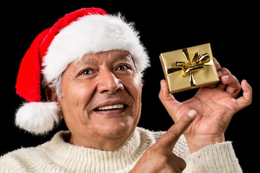 Smiling old man with challenging gaze is gesturing with his raised right index finger. He is pointing at his left hand holding a small, golden, wrapped present. Red Father Christmas cap. Ample DOF.