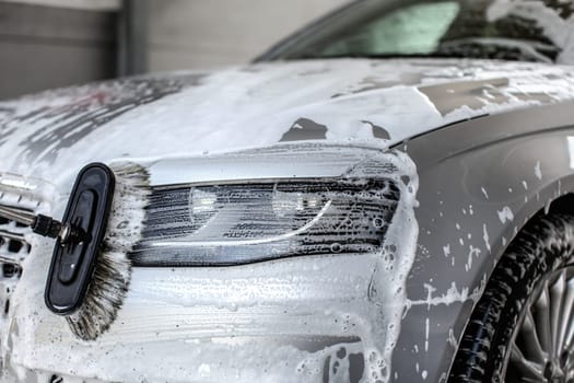 Front light of silver car, covered with shampoo and foam, being washed in manual carwash with brush.