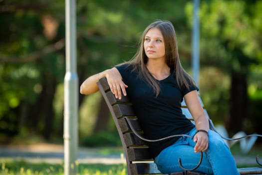 A beautiful girl in casual clothes sits on a bench in a beautiful green park