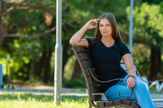A beautiful girl in casual clothes sits on a bench in a beautiful green park, the girl looks into the frame