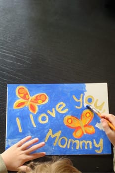 hand of a little child painting a picture with butterflies for mommy at mother's day with the text I love you mommy