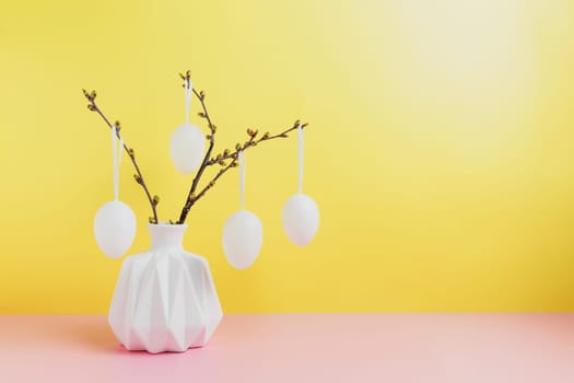 Happy Easter holiday, coming spring concept. Tree branches, flower twigs in white vase or jar with hanging eggs. Yellow and pink table background with copy space. Easter preparation, greeting card