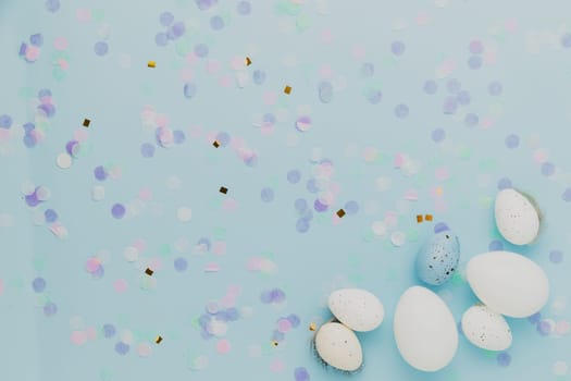 Happy Easter holiday, coming spring concept. Flat lay with white, colorful quail eggs, feather. Blue table background, paper confetti, copy space for ad or text. Easter greeting card, invitation