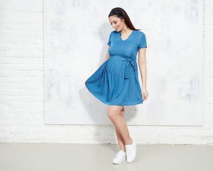 Fashion portrait of happy pregnant woman in blue summer dress against white wall with copyspace.