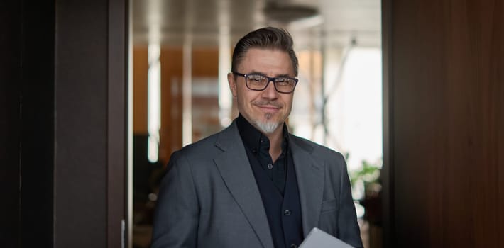 Business portrait - confident businessman standing in office. Happy mid adult man in shirt and jacket, smiling. Bearded, gray hair.