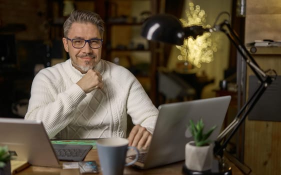 Happy mid adult man working with laptop computer in home office, glasses, gray hair. Winter, Christmas, dark cosy room.