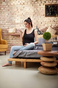 Happy pregnant woman sitting at home smiling, relaxing using mobile phone.