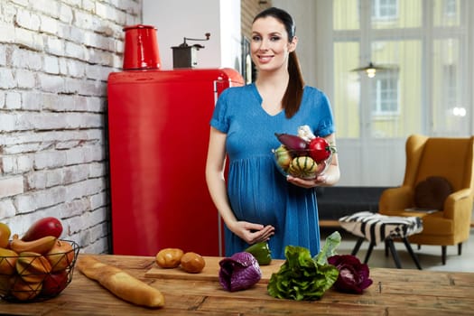 Happy pregnant woman at home in kitchen with living room in background preparing salad, eating healthy food from vegetables.