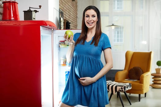 Happy pregnant woman at home in kitchen with living room in background taking healthy food from fridge, eating green apple.