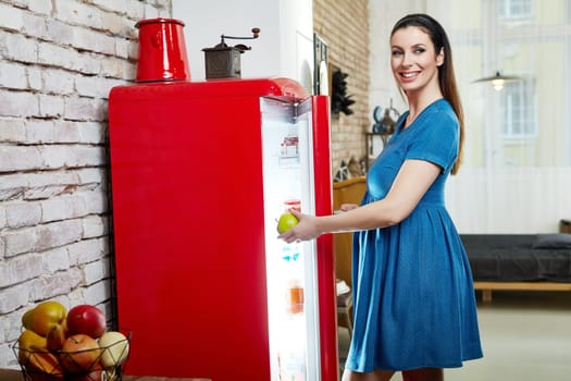 Happy pregnant woman at home in kitchen with living room in background taking healthy food from fridge, eating green apple.
