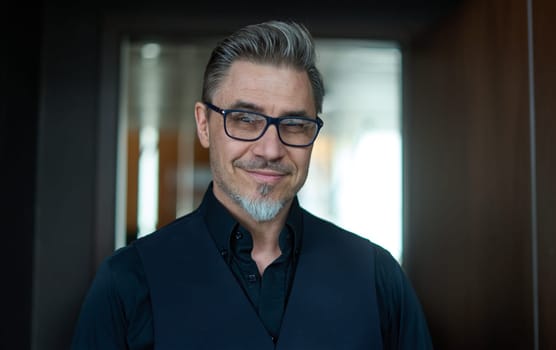 Business portrait - businessman standing in office, smiling. Happy middle aged, mid adult man in black jacket.