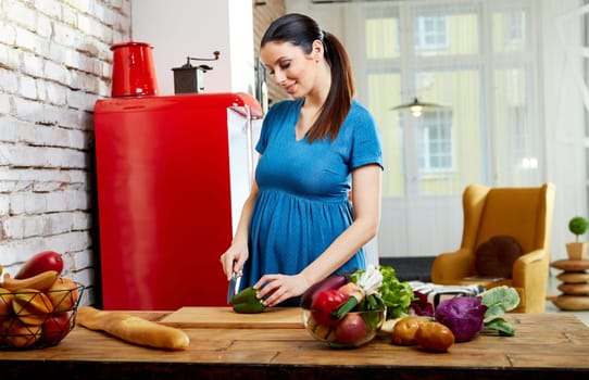 Happy pregnant woman at home in kitchen with living room in background preparing salad, eating healthy food from vegetables.