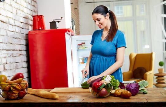 Happy pregnant woman at home in kitchen with living room in background preparing salad, eating healthy food from vegetables.