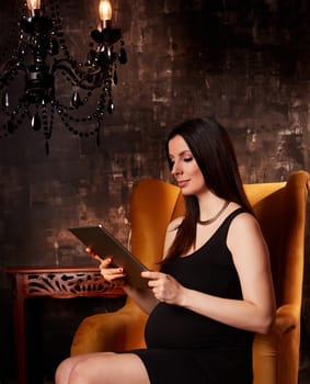 Beautiful pregnant woman in elegant black dress sitting in hotel lobby holding handheld tablet.
