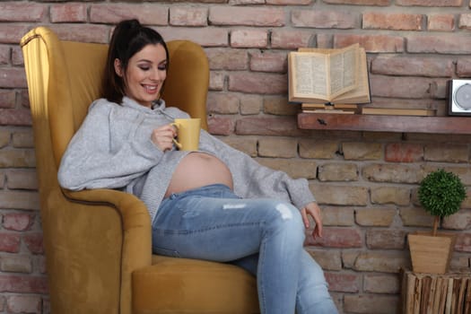 Happy pregnant woman at home relaxing in armchair holding cup of tea.