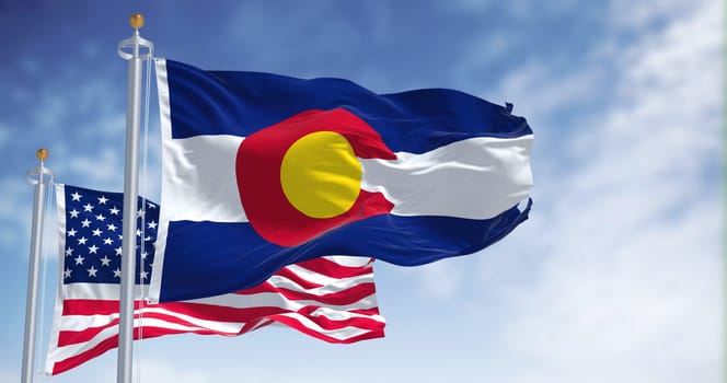 The Colorado state flag waving along with the national flag of the United States of America. In the background there is a clear sky.