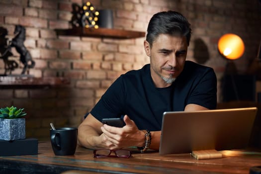 Older white man sitting at desk in dark room at home, working, using phone and tablet
