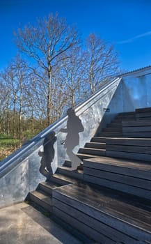 unknown young woman and her shadow walking down wooden stairs (metall contruction) in an outdoor leisure area, bright weather and blue sky, digitally processed