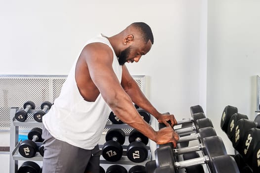 An African American man stands in front of a mirror and leans on dumbbells in the gym. High quality photo