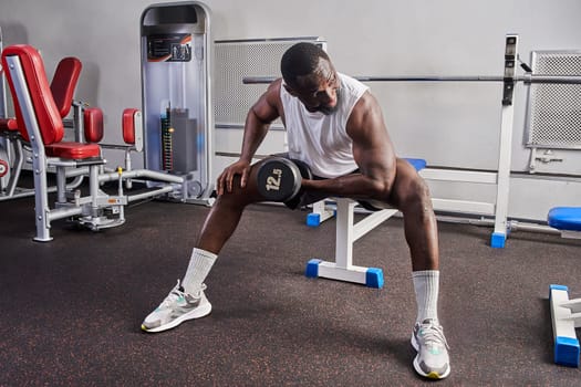 An African American man shakes his biceps while sitting on a bench in the gym. High quality photo