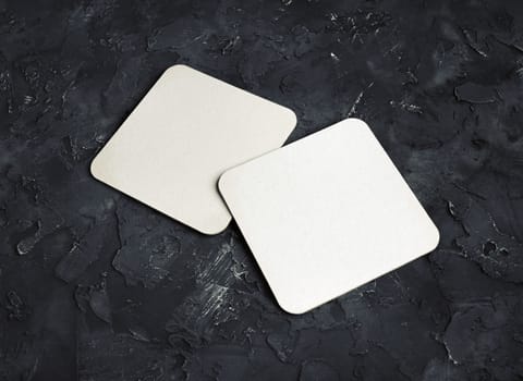 Blank white square beer coasters on black plaster background.