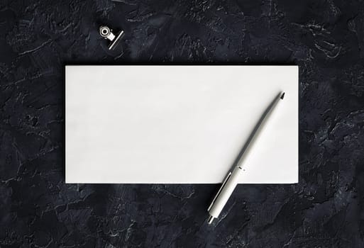 Blank envelope, pen and clip on black plaster background. Branding mock up. Top view.