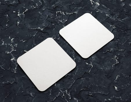 Two blank square beer coasters on black plaster background.