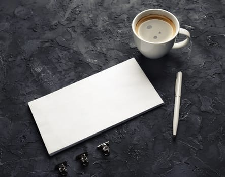 Photo of blank paper envelope, coffee cup, pen and metal clips on black plaster background. Blank stationery set.