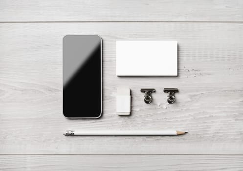 Smartphone and blank stationery: business card, pencil, eraser and clips on light wooden background. Flat lay.