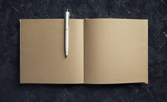 Open blank craft paper sketchbook and pen on black plaster background. Branding mock up.