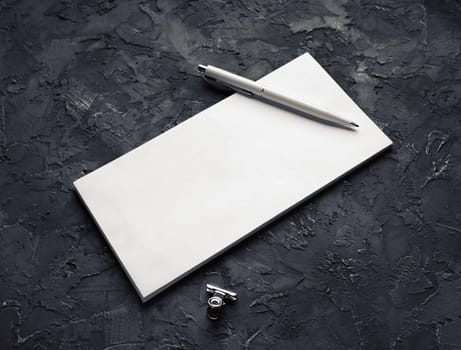 Photo of blank paper envelope, pen and metal clip on black plaster background.