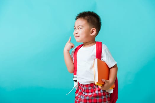 Back to school concept. Portrait Asian happy funny cute little child boy smile hug books and point finger to side away space, isolated blue background. Kid from preschool kindergarten with school bag