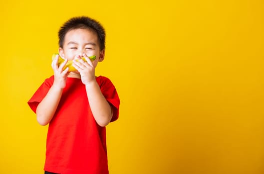 Happy portrait Asian child or kid cute little boy attractive smile wearing red t-shirt playing holds banana fruit, studio shot isolated on yellow background