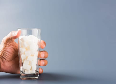 Hand hold on glass full of white sugar cube sweet food ingredient, isolated on gray background, health high blood risk of diabetes and calorie intake concept and unhealthy drink