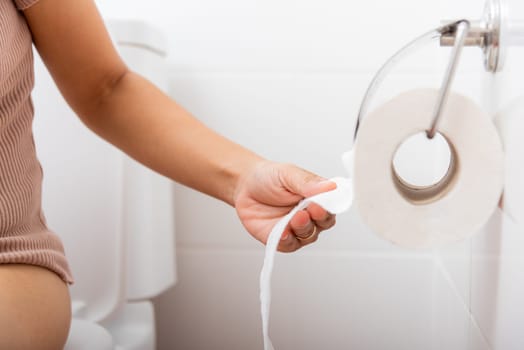Closeup hand pulling toilet paper roll in holder for wipe, woman sitting on toilet she taking and tearing white tissue on wall to towel clean in bathroom, Healthcare concept