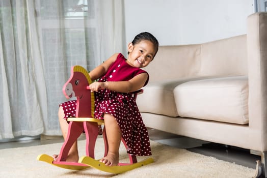 Asian cute little girl swinging riding toy horse, Smiling kid playing horse rocking chair at home in living room, Happy time to play during self kindergarten preschool