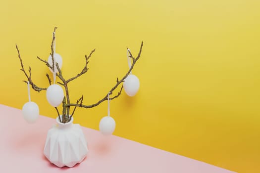 Happy Easter holiday, coming spring concept. Tree branches, flower twigs in white vase or jar with hanging eggs. Yellow and pink table background with copy space for ad or text. Easter home decoration