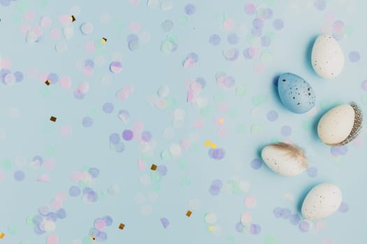Happy Easter holiday, coming spring concept. Flat lay with white, colorful quail eggs, feather. Blue table background, paper confetti, copy space for ad or text. Easter greeting card, invitation