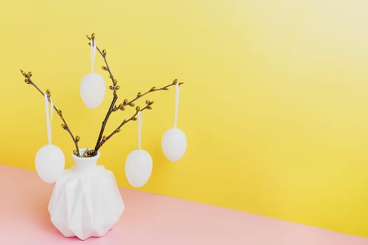 Happy Easter holiday, coming spring concept. Tree branches, flower twigs in white vase or jar with hanging eggs. Yellow and pink table background with copy space. Easter preparation, greeting card