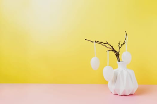Happy Easter holiday, coming spring concept. Tree branches, flower twigs in white vase or jar with hanging eggs. Yellow and pink table background with copy space. Easter preparation, greeting card